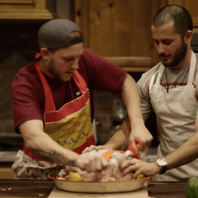 Two men in aprons preparing raw chicken in a kitchen setting.